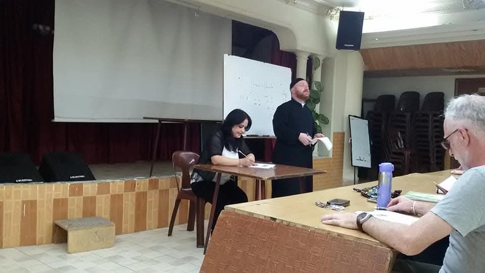 A priest and a coordinator at the front of the hall beside a whiteboard with Syriac writing