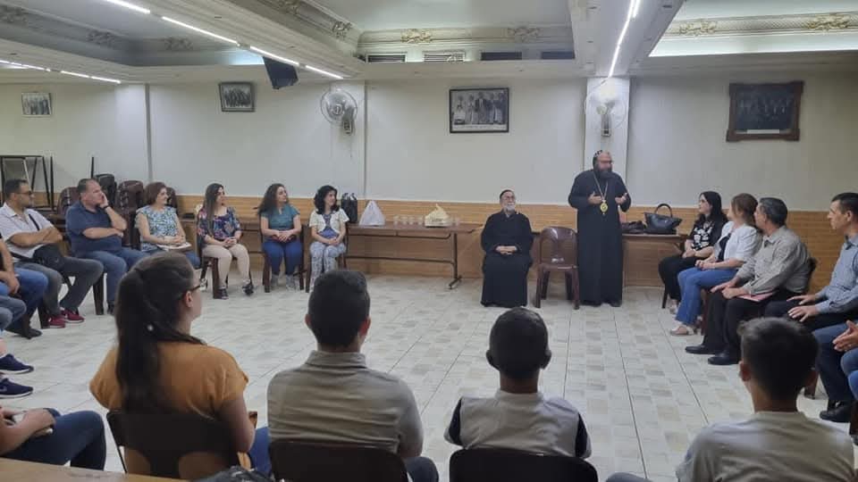 Young people seated in a circle listening to a priest speaking in the association hall