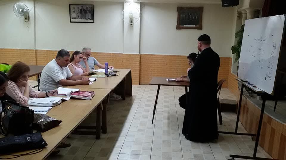 Learners seated around long study tables with books open while a teacher listens to a reader