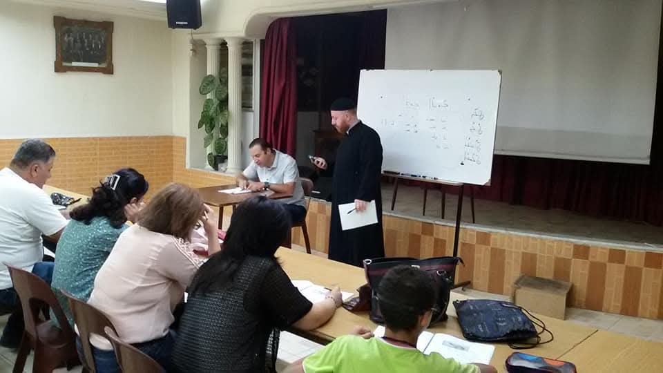 A teacher standing beside a whiteboard covered in Syriac script while learners take notes