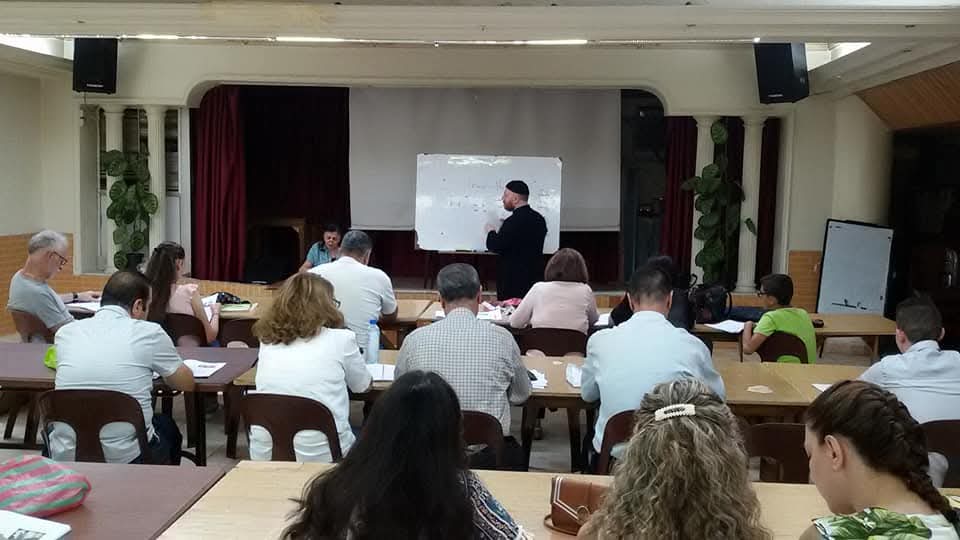 Learners at long tables facing a teacher at a whiteboard in the association hall