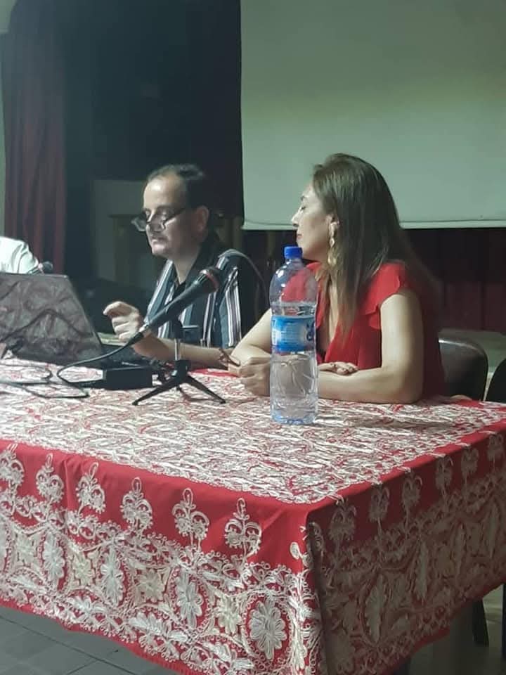 Two speakers seated behind microphones and a laptop during a cultural lecture