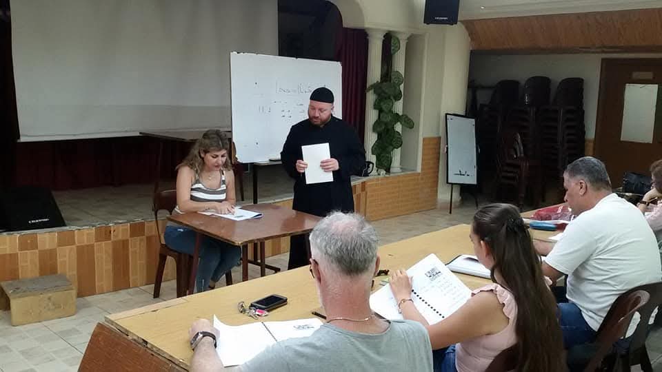 A priest reading from a sheet beside a coordinator at a table while participants take notes