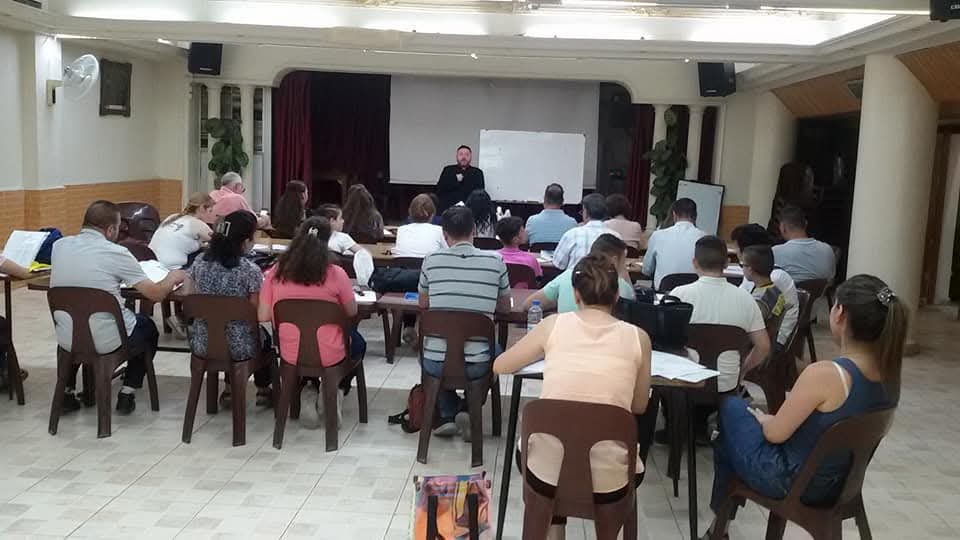 Adults and children seated at tables facing a teacher and whiteboard during a Syriac language class